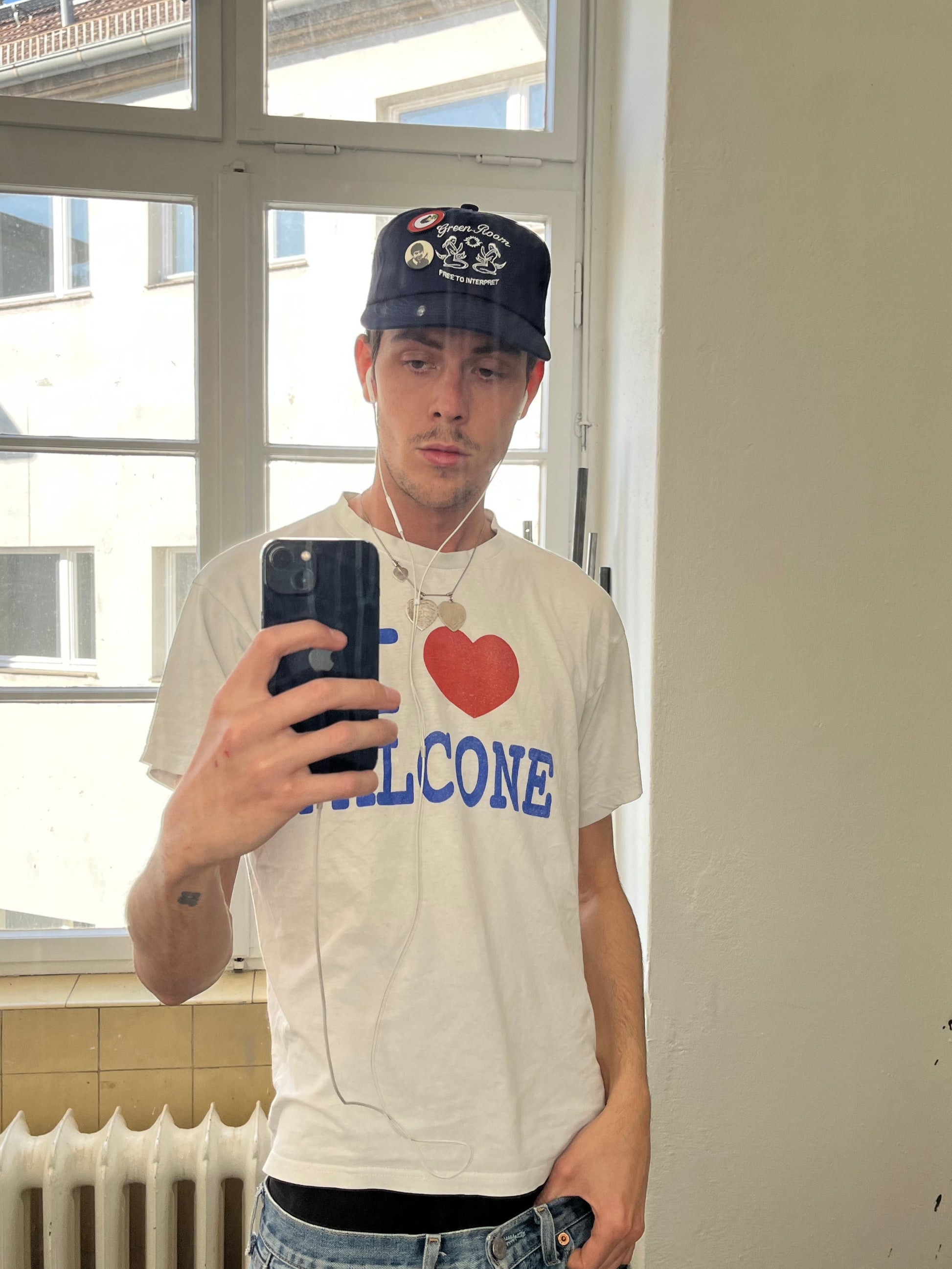 Person trägt eine navyfarbene Cap mit weißer Meerjungfrauen-Stickerei und „Green Room“-Schriftzug auf der Vorderseite. Die Cap ist mit Buttons verziert. Dazu wird ein weißes T-Shirt mit „I ❤️ FALCONE“-Druck getragen.