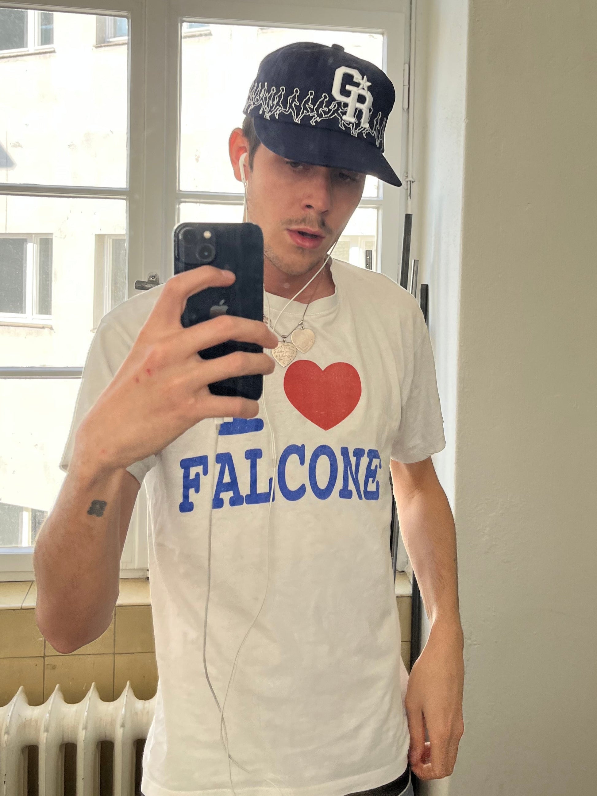 Selfie eines Models mit weißem T-Shirt und navyblauer Cap mit „GR“-Logo und umlaufender Stickerei weißer Laufsilhouetten, aufgenommen vor einem Fenster.