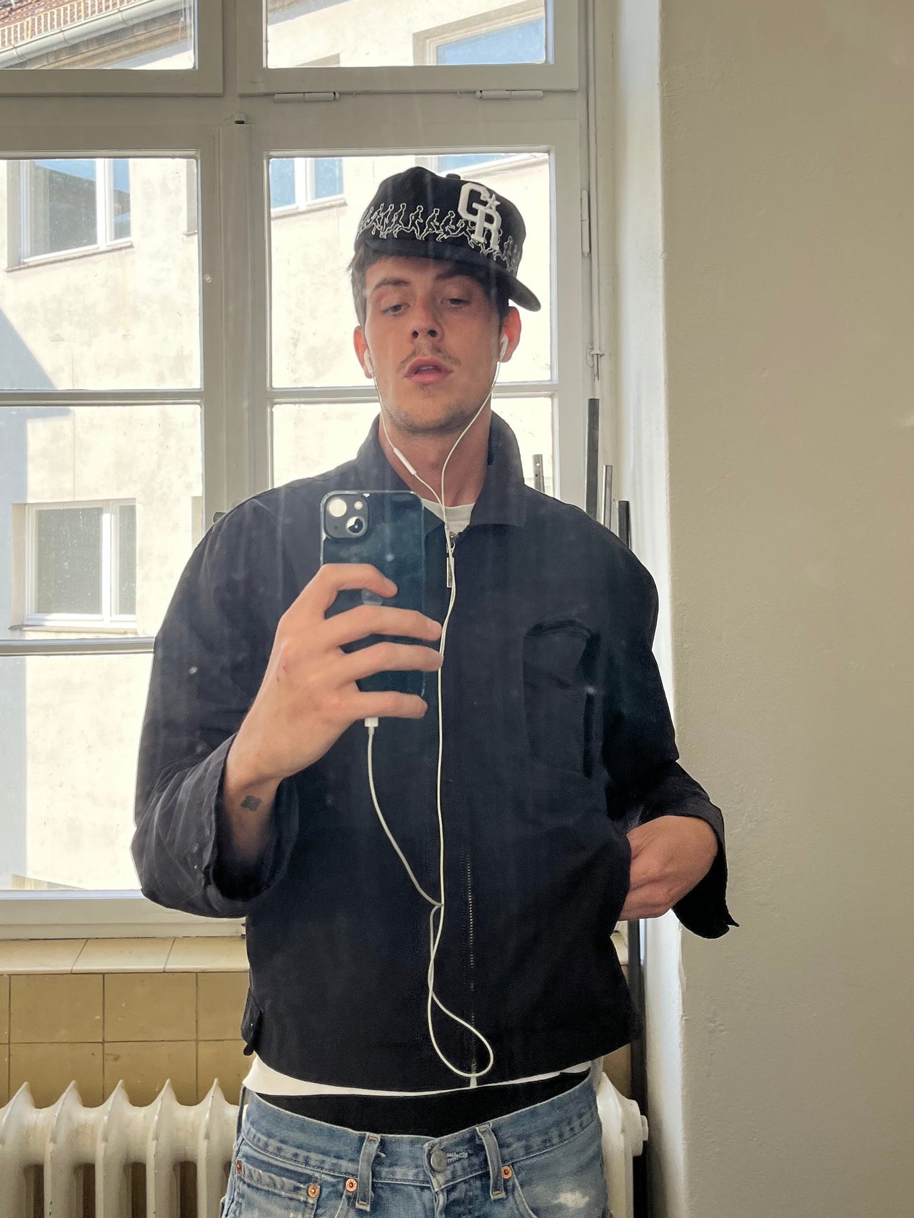 Selfie eines Models mit dunkler Jacke und schwarzer Kappe mit „GR“-Logo und umlaufender Stickerei weißer Laufsilhouetten, aufgenommen vor einem Fenster.