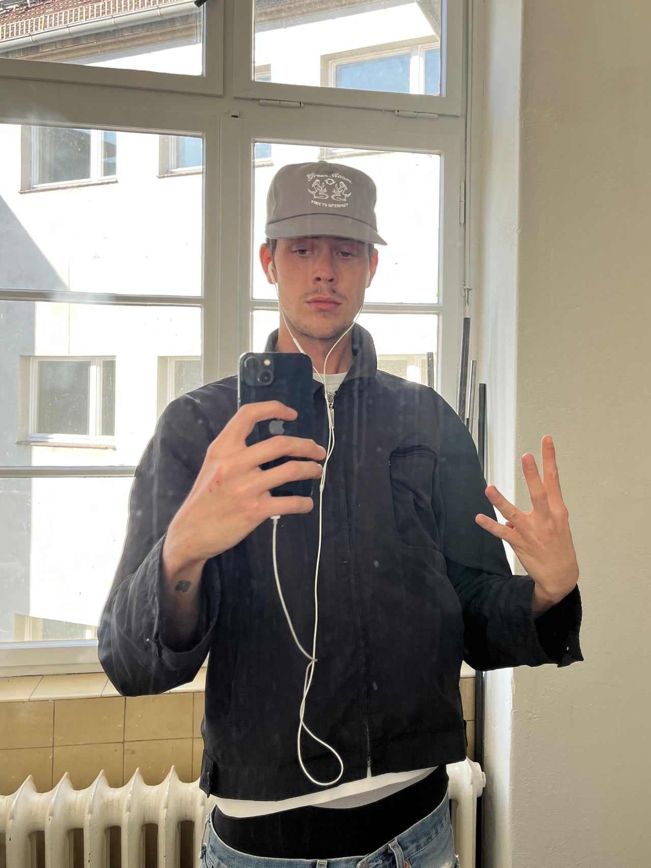 Person in dunkler Jacke und hellem T-Shirt trägt die graue Green-Room-Cap mit Stickmotiv vorne. Die Cap sitzt gerade auf dem Kopf und das Motiv mit Meerjungfrauen und Schriftzug ist deutlich zu erkennen.